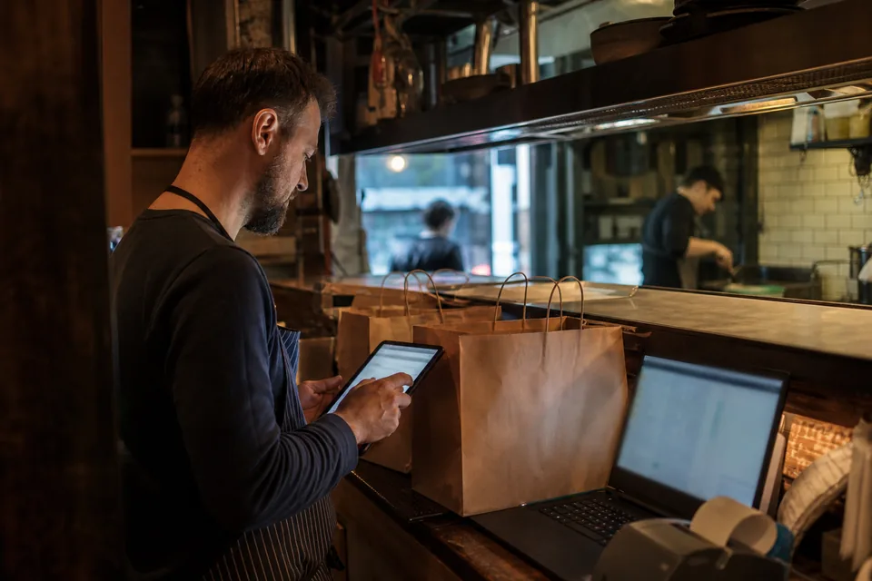 Właściciel bistro sprawdza na tablecie zamówienia z dowozem obok toreb na wynos i laptopa z rozliczeniem
